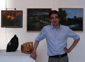 Man with shirt sleeves rolled up beside two small sculptures wtih three medium sized paintings on the wall behind him