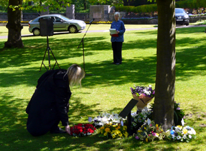 Woman in black crouching to lay a wreath among others at the foot of the memorial tree