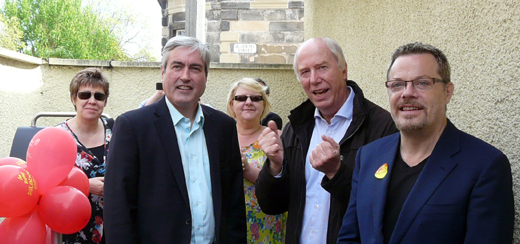 Lesley in black floral dress and shades, Iain in suit without tie, Karen in yellow patterned frock with shades, Malcolm in anorak cheering and Eddie in blue suit, smiling