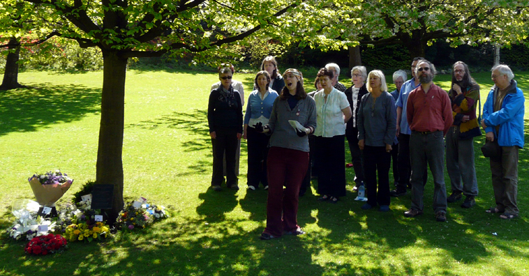 Group of a dozen people singing under a tree in Princes St Gardens, which has bouquets of flowers laid around it