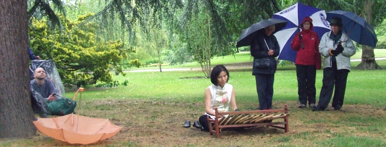 Seated woman plays a percussion instrument of bamboo pipes under a coniferous tree, as three women stand beneath their umbrellas watching on