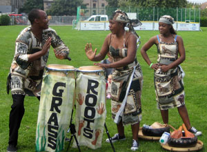 Man and woman playing drums, with the travelling five a side football pitch in the background