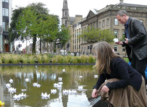 Fay Young kneeling and Gordon Munro standing cast white paper lotuses on the pond
