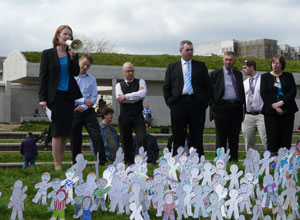 A woman MSP addresses the lobbiers with the grass bank of the Parliament in the background