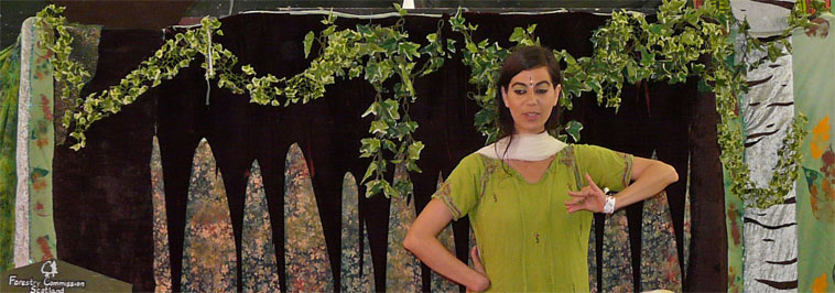 A woman in olive green telling a story with her hands  against a decorated forest background