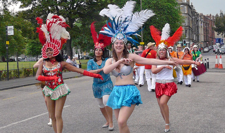 Scarlet and blue clad dancers with golden harp and cadduceus, adorned with gold feathers and a crimson garbed young woman smiling in the middle