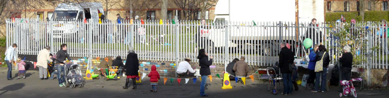 Long view of the aluminum fence between the play park and the grassy area with children and parents painting bricks along itwith 