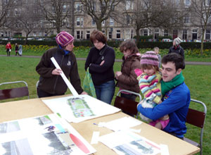 A woman discusses the plan with a young family which sits round some tables in the park