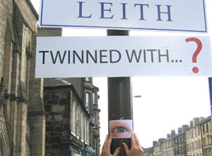 Signs: LEITH & TWINNED WITH...? with artist Gavin Lockhart;s hands placing a picture of an eye on the lamppost