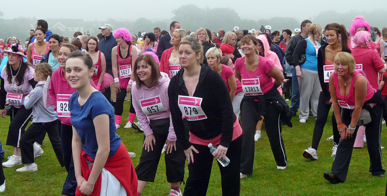 Around ten women in the foreground stretching their hamstrings, with others visible behind. Most of them are smiling