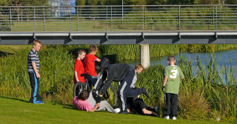 Boys looking in the reeds at the edge of a pond with a metal walkway above them