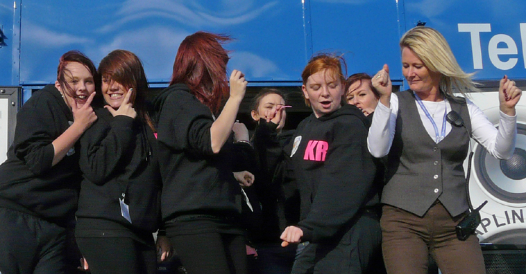 Girls in black sweat shirts and jeans dancing with a woman in grey waistcoat and slacks