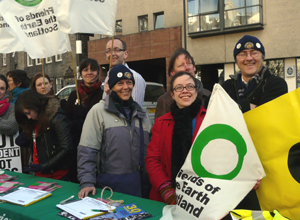 People smiling behind a pasting table, holding Friends of the Earth Scotland flags