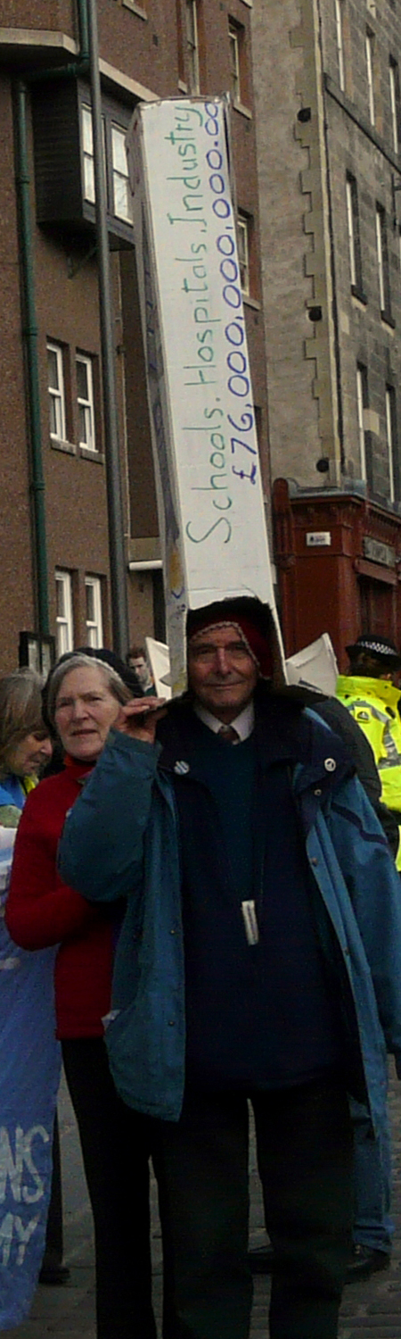 Man with a tall home made cardboard hat which reads vertically "Schools, Hospital, Industry £76,000,000,000.00