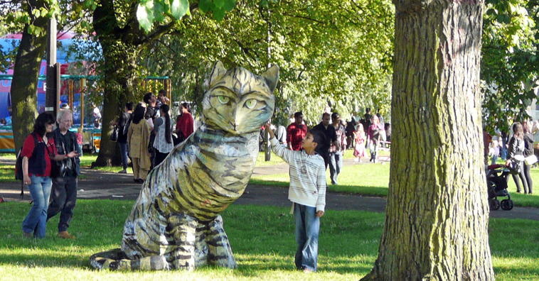 A boy strokes the giant papier mache tabby cat under the trees od Leith Links as visitors pass by