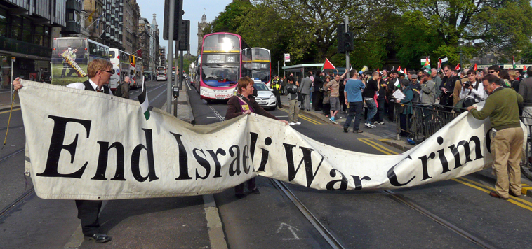 "End Israeli War Crimes" banner being organised with buses behind, while man with loud hailer explains what is happening