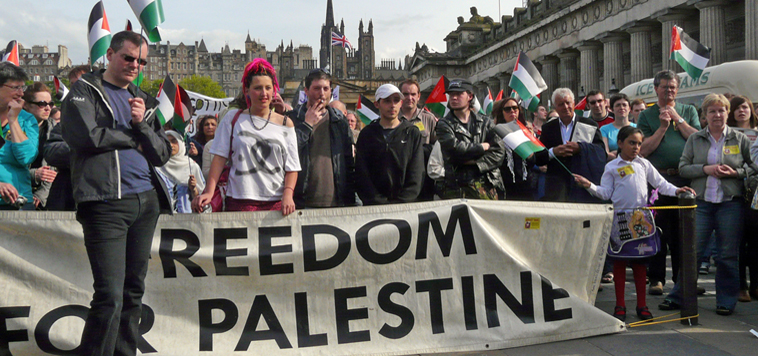 People lined behind a banner "Freedom for Palestine" , waving Palestinian flags. The National Galleries Mound Complex is in the background