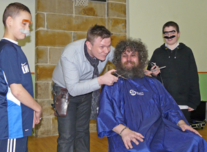 Simon holding his scissors near Dave's throat as two boys in false moustaches watch on