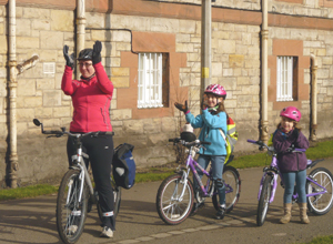 Woman in red on bicycle applauds the march as two girls on bikes cheer on.  All three are wearing cycle helmets