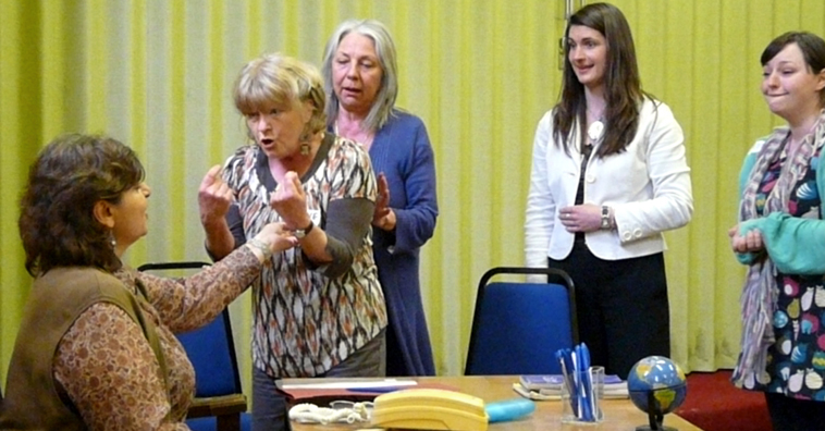 Angry woman stands and gesticulates at the seated Caroline as Penny tries to calm her and to other women watch on