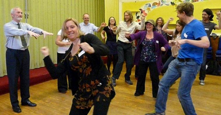 People dancing around a man in shirt and tie who is standing with his arms extended in front of him