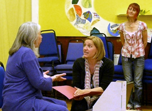 Seated woman with palms outstretched talking to a crouching woman with an open pink folder