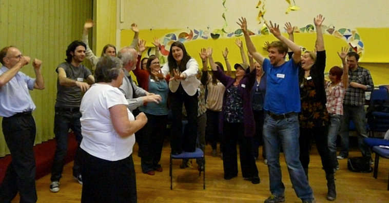 Woman standing on chair with arms outstretched exhorts woman and man surrounded by group with raised arms