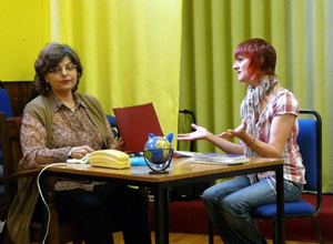 Woman seated behind a desk, ignoring a woman seated in front of her desk with palms outstretcched