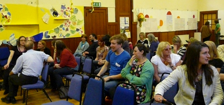 Around 20 people seated on blue chairs sharing ideas