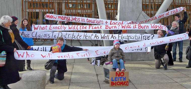 Zoe sits in front of banners asking questions about the Leith Biomass Plant such as "Who will verify Forth Port' claim of carbon savings?