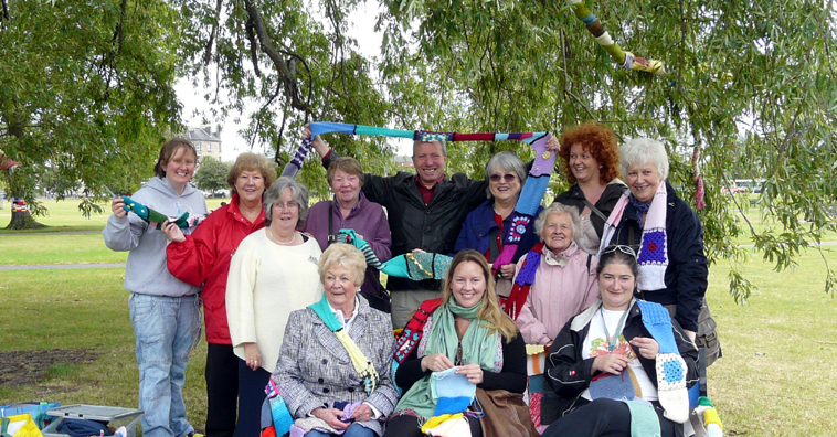 Group of women seated on a bench beneath a willow tree on Leith Links with women standing behind them, as Leith Cllr Gordon Munro grins, holding a scarf aloft
