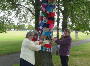 Three smiling women wrapping woolen scarves round a tree