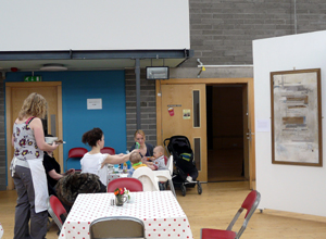 A woman serves mothers and small children at a table with Edwin Muir's "GGLC" on display behind them