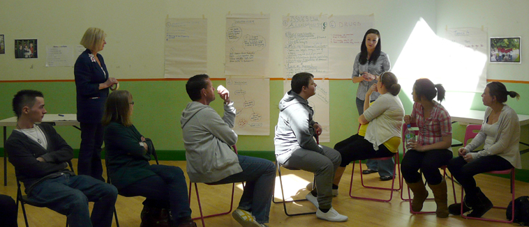 A young woman standing by flip chart pages listens to a group of young people as Maggie Havergal in a casual blue suit watches on
