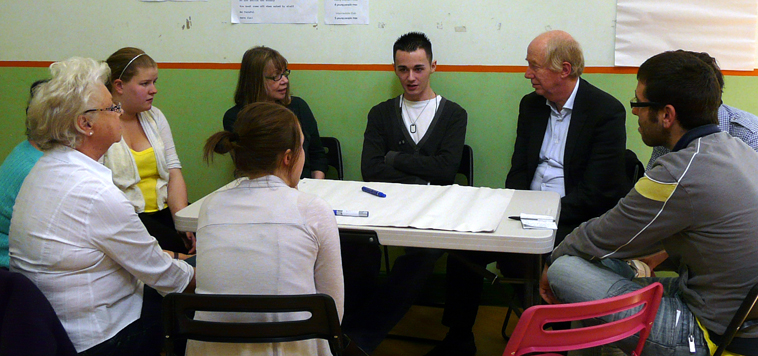 Group of people seated round a table listening to a young man talking