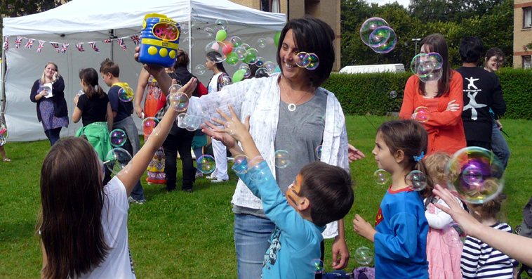 Smiling woman holds up bubbling bubble machine as children reach up to the the bubbles