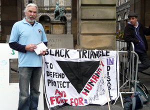 Bearded man in a sky blue rugby shirt, stands beside a banner: BLACK TRIANGLE TO DEFEND PROTECT AND FIGHT FOR HUMANITY IN DISABILITY