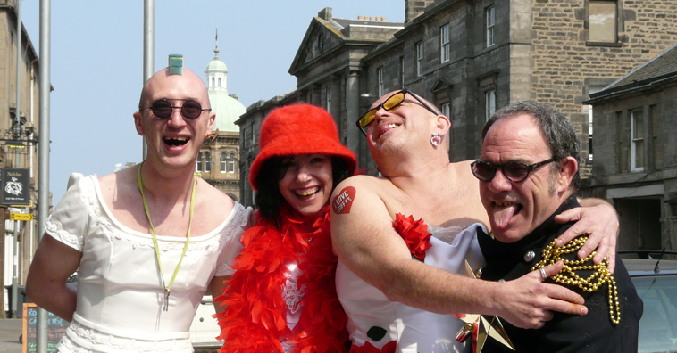 Man in bridal dress with Mohican made of washing up sponges, woman in white with red accessories, man in wedding dress, and man in black uniform with gold braid sticking his tongue out 