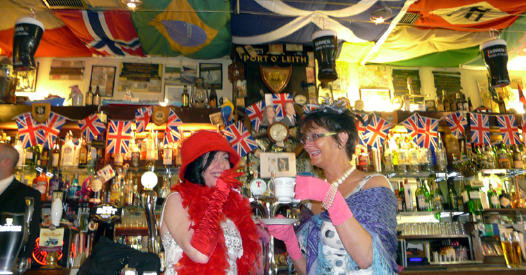 Woman in a white dress with red felt hat, feather boa and cigarette lighter and woman in white dress with pink gloves, blue shawl and teacup