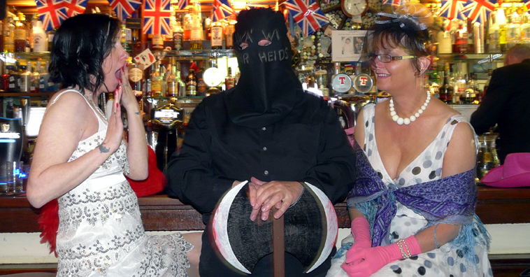 Two women in white dresses sit at a bar staring in horror and bemusement at a man dressed in black as an Executioner resting his hands on an axe 