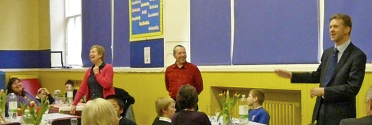 The three Councillors stand to speak to the school in the gym hall, with Mrs Thomson and pupils seated at dining tables