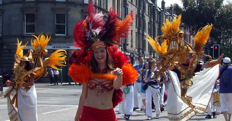 White clad dancers with golden harp and cadduceus, adorned with gold feathers and a crimson garbed young woman smiling in the middle