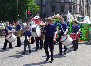 Young men in blue t-shirts march with drums in front of the yellow Citadel Youth Centre banner